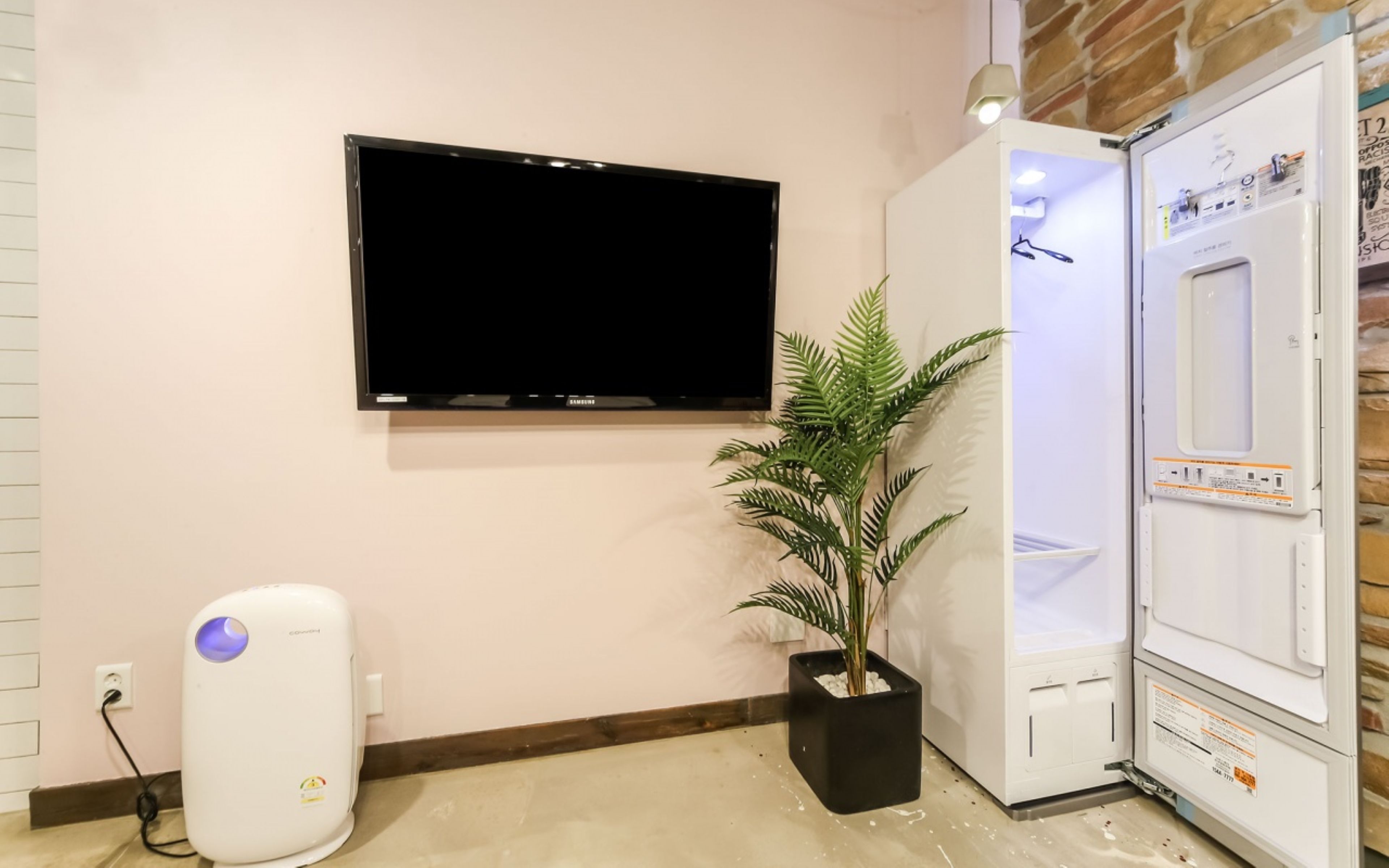A room with a flat-screen TV mounted on the wall, a white air purifier, a potted plant, and a white appliance with a control panel.