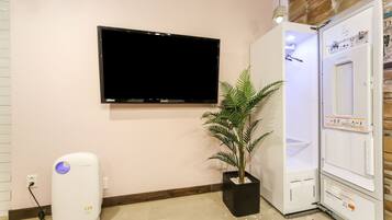 A room with a flat-screen TV mounted on the wall, a white air purifier, a potted plant, and a white appliance with a control panel.