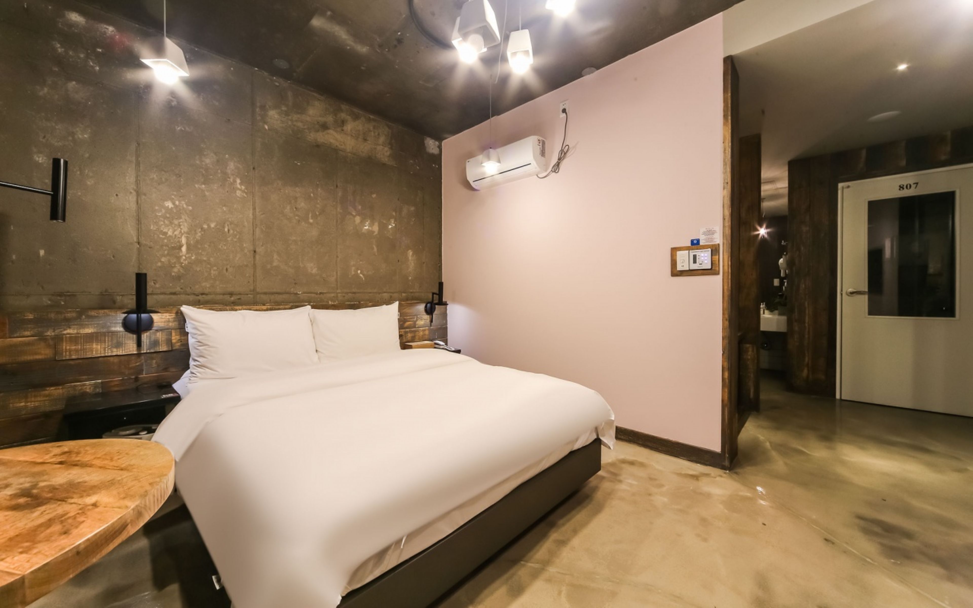 A modern hotel room with a large bed, a wooden headboard, a round wooden table, and a built-in air conditioning unit.