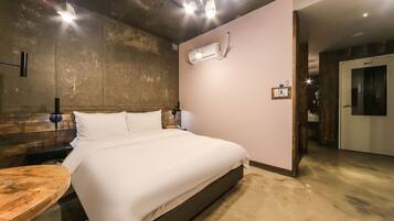 A modern hotel room with a large bed, a wooden headboard, a round wooden table, and a built-in air conditioning unit.