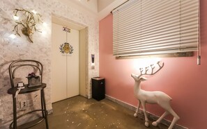Un couloir d’hôtel avec une porte blanche, une statue de cerf, un luminaire fixé au mur et un présentoir en métal sur lequel est posée une plante.