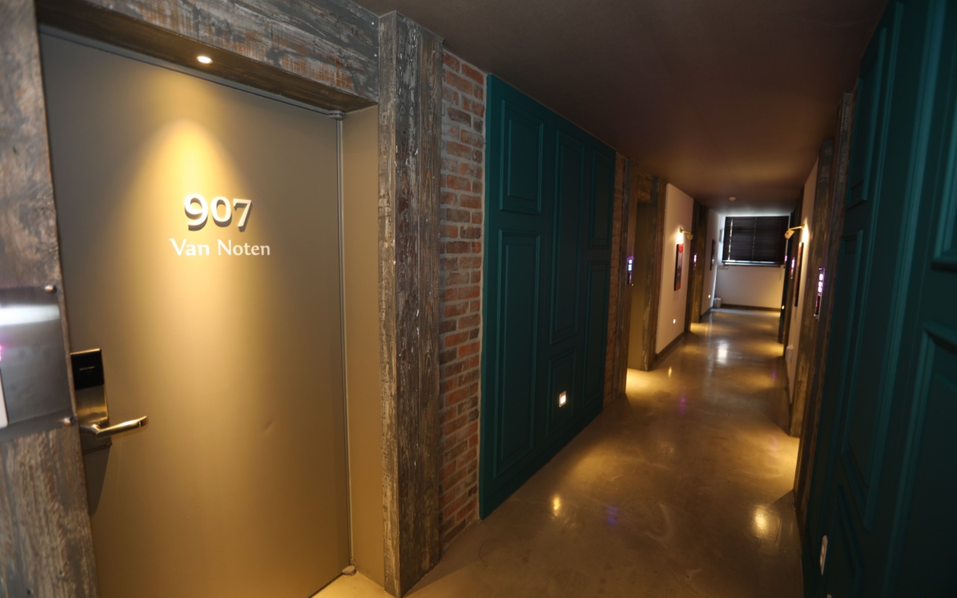 Een gang met een bordje met de tekst '907 Van Noten' aan de linkerkant, die leidt naar een kamer met een bakstenen muur en blauwgroene deuren.