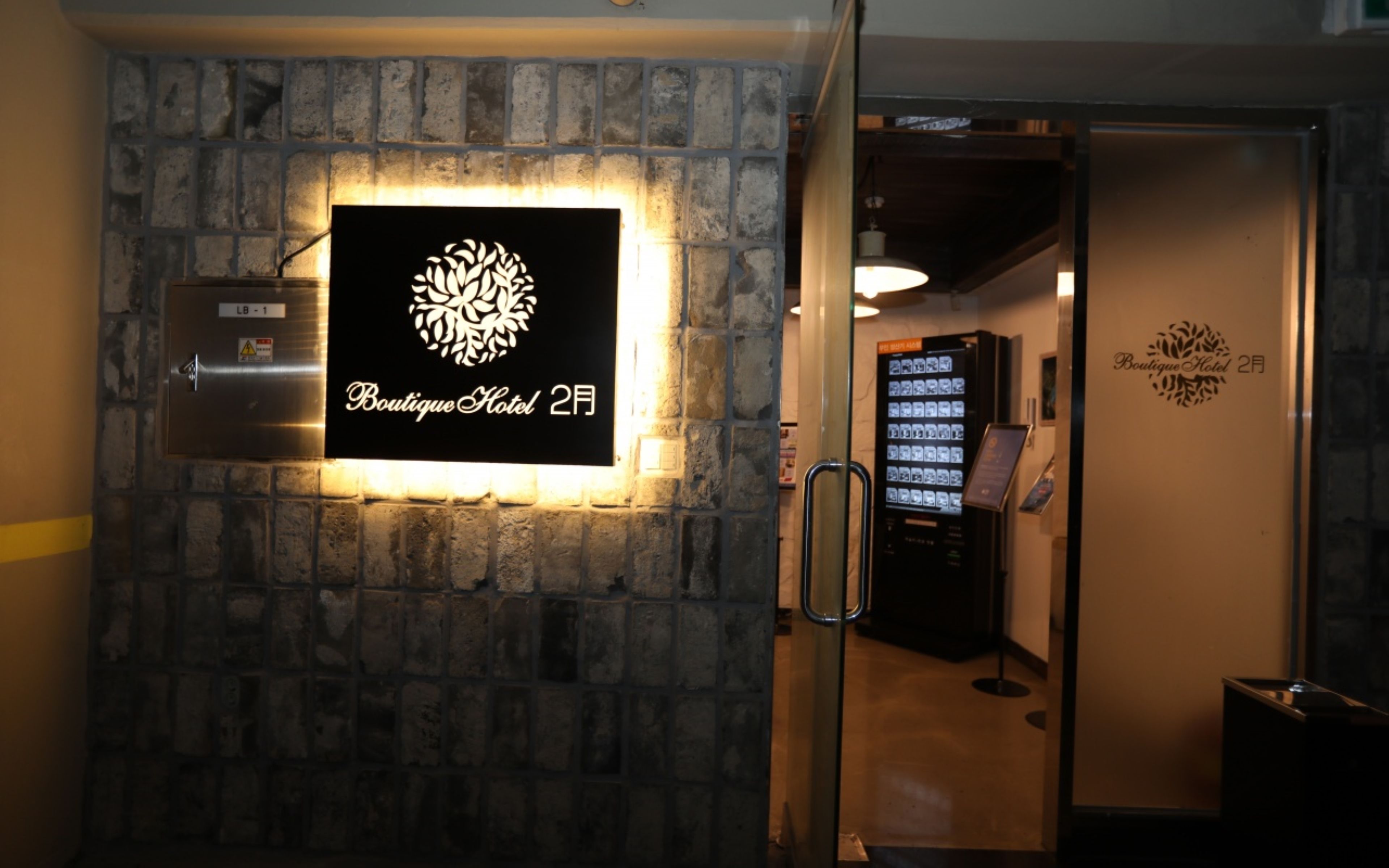 Eine an der Wand befestigte Schilder für das Boutique Hotel 2F mit einem dekorativen Wappen, ein Getränkeautomaten und ein Glastür-Eingang.