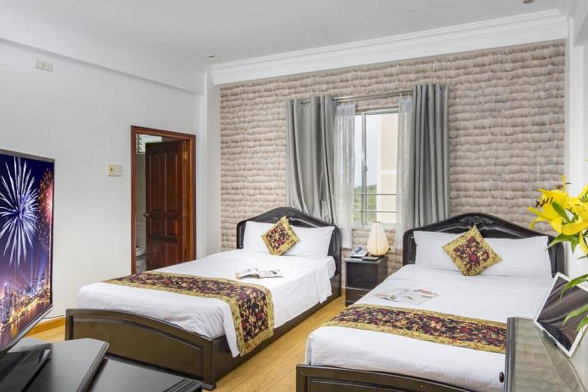 A hotel room with two beds, a large window with curtains, a wooden door, and a wall-mounted TV.