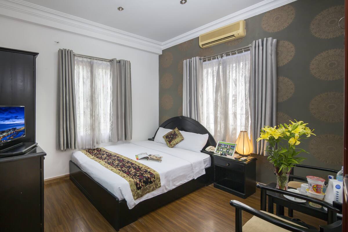A hotel room with a bed, a desk, a chair, a TV, a lamp, and a vase with flowers.