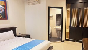 Una habitación de hotel con cama, mesita de noche, equipo de aire acondicionado, baño con lavabo e inodoro, y armario con puerta corredera.