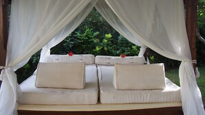 Una cama con dosel, con ropa de cama y almohadas blancas, ubicada en una estructura de madera con vista a la vegetación.