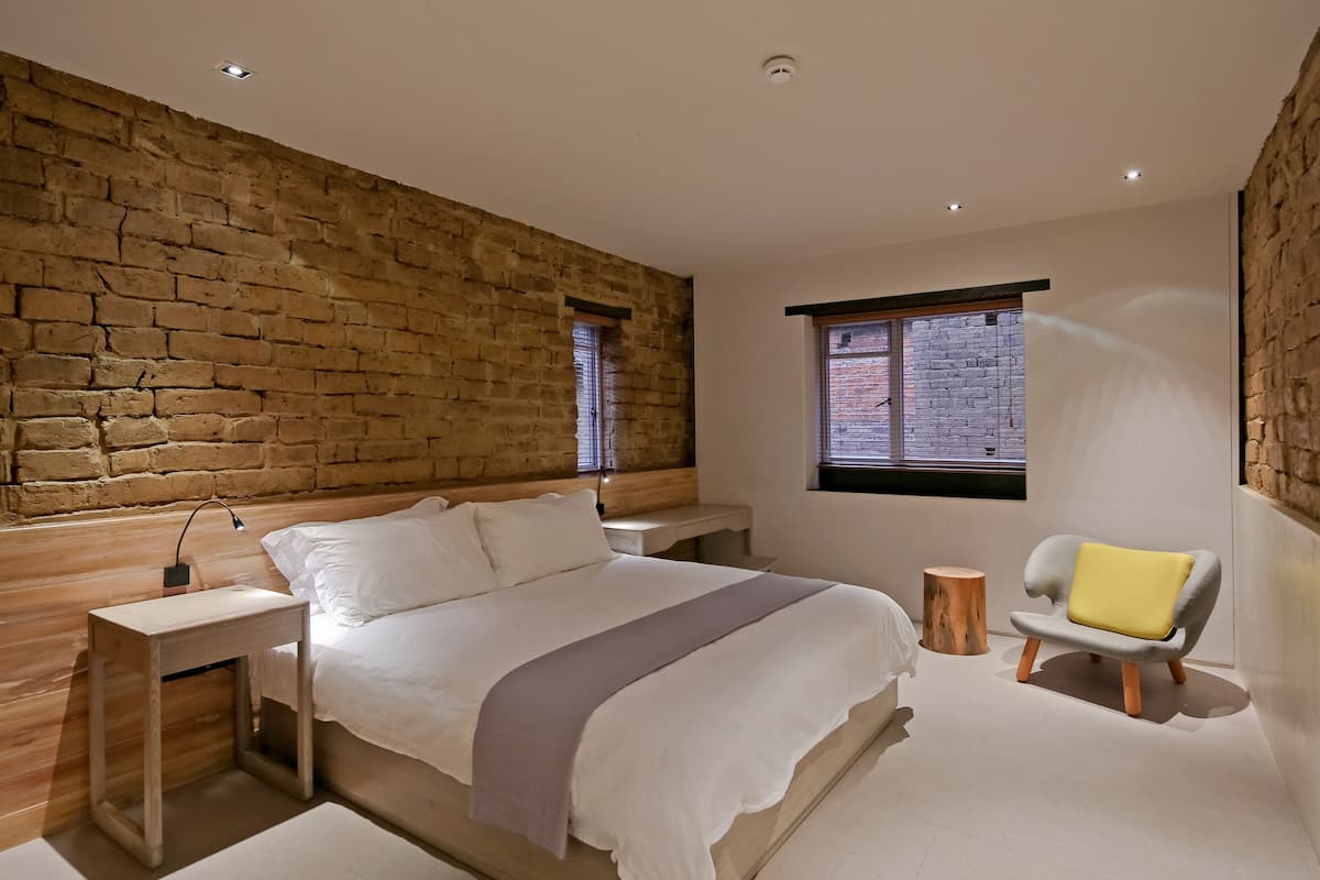 A modern bedroom with a stone wall, a large bed, a bedside table, a chair, and a window with a view.
