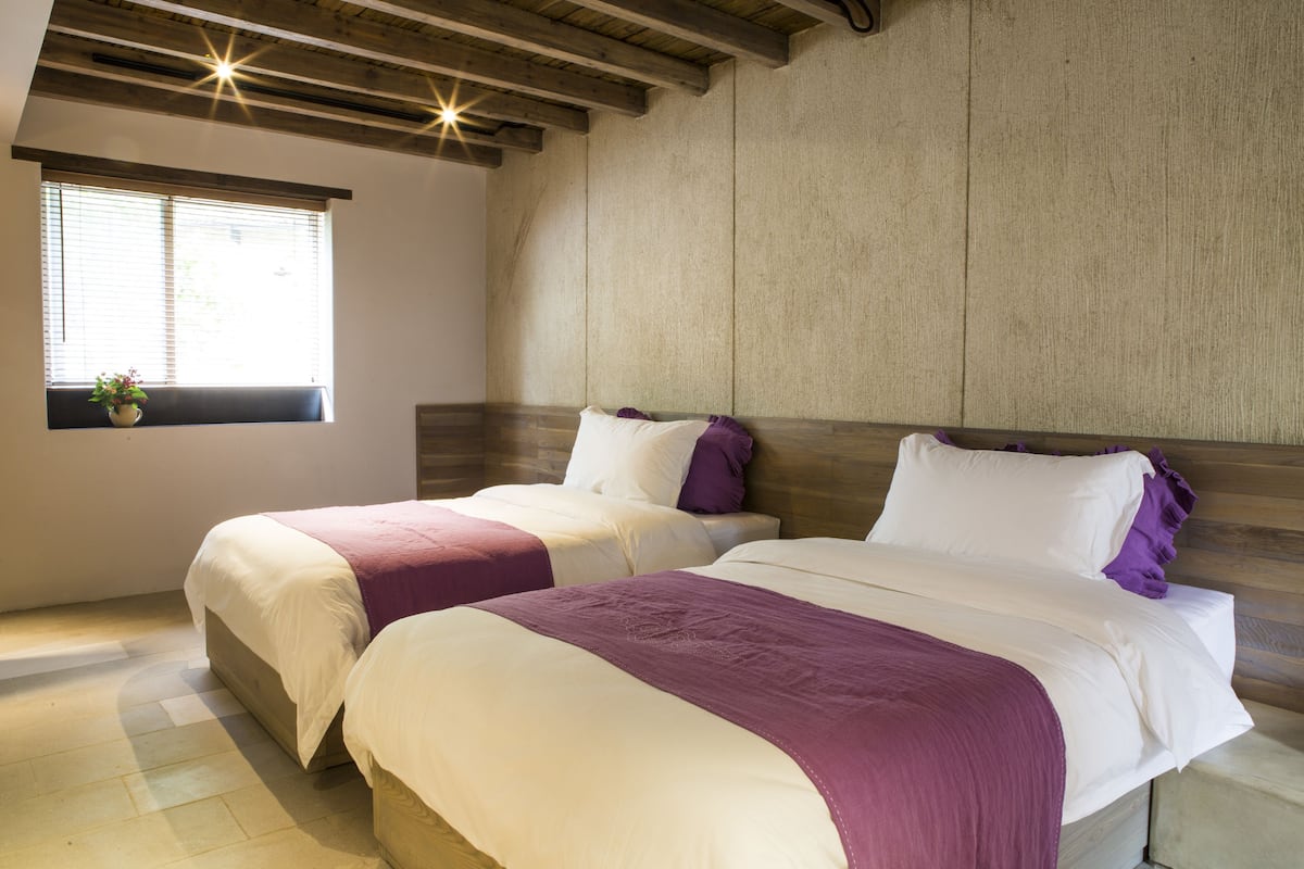 A bedroom with two beds, a window with blinds, and a wooden ceiling.