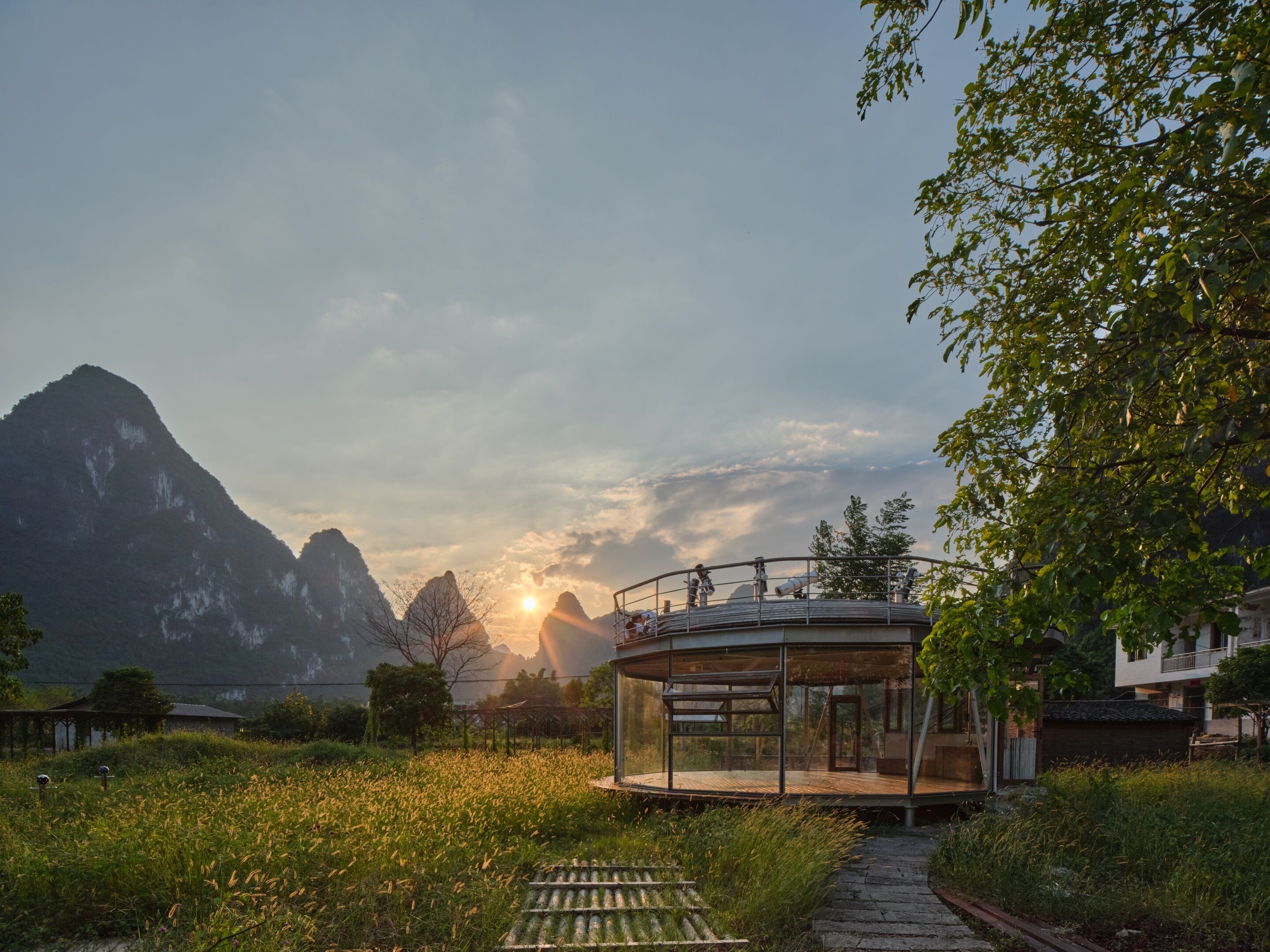 A modern glass house with a panoramic view of mountains and a sunset.
