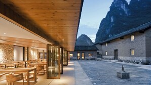 A modern building with a wooden ceiling and stone walls, featuring a courtyard with tables and chairs, and a view of mountains in the background. - XY YunHouse (Guilin)