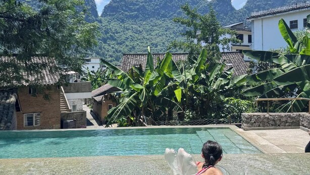 A clear pool with a person in the water, surrounded by lush greenery and traditional buildings. - XY YunHouse (Guilin)