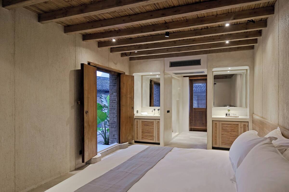 A bedroom with a large bed, two wooden cabinets, a mirror, and a door leading to a balcony with a view of greenery.