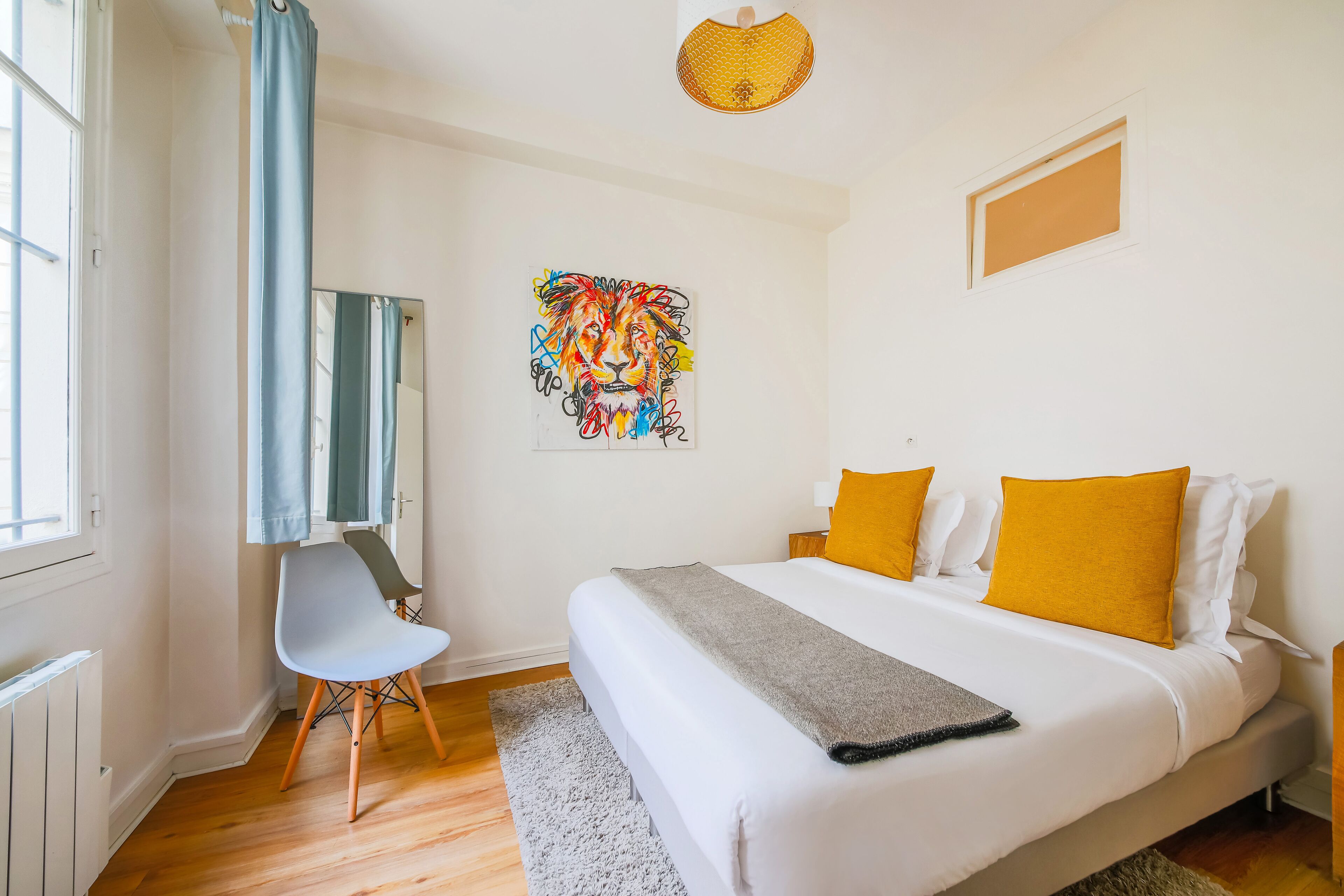 A bedroom with a bed, two pillows, a chair, a desk, a mirror, a wall art piece, and a radiator.