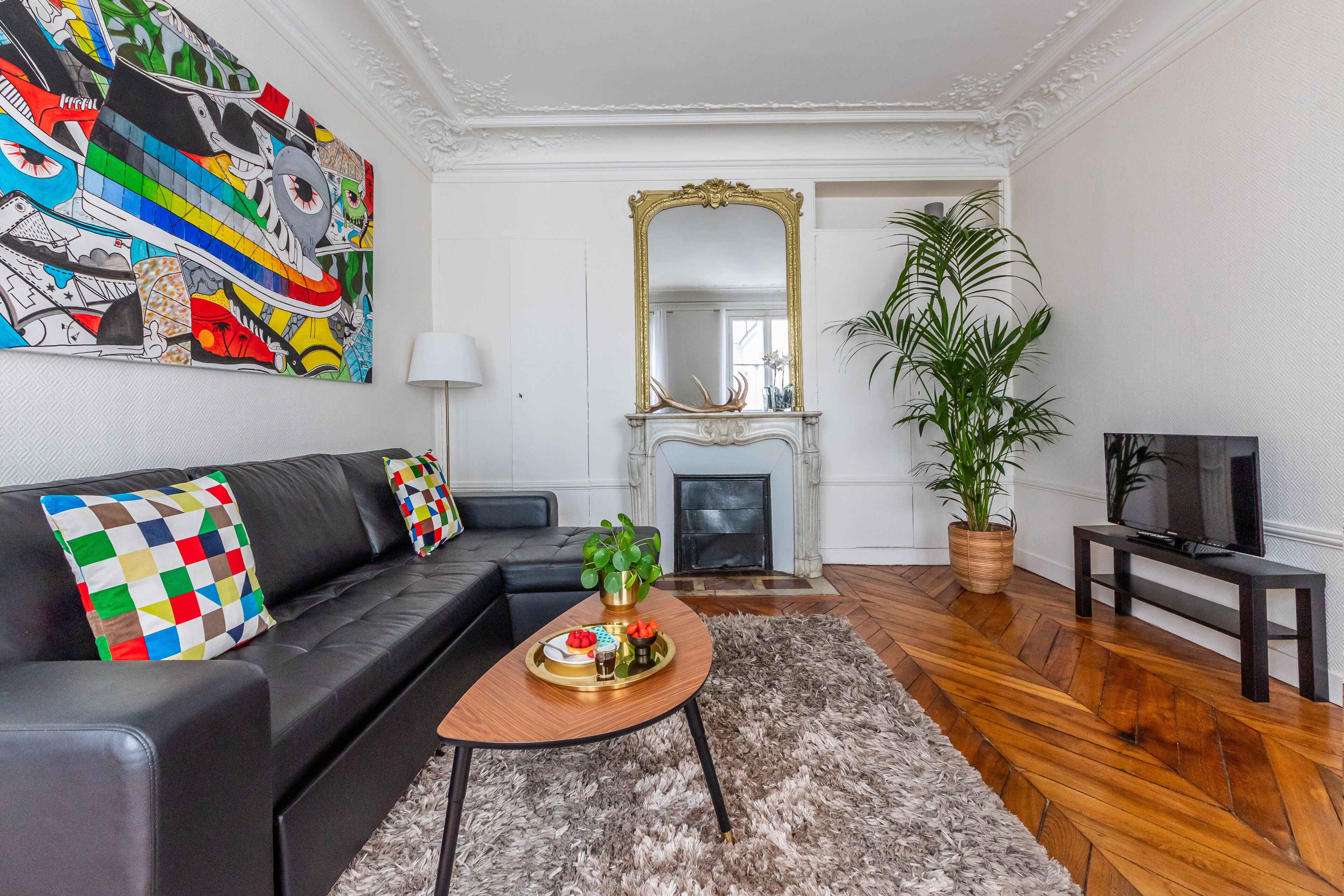 A modern living room with a black leather sofa, a wooden coffee table, a colorful abstract painting, a large mirror, a fireplace, and a television.