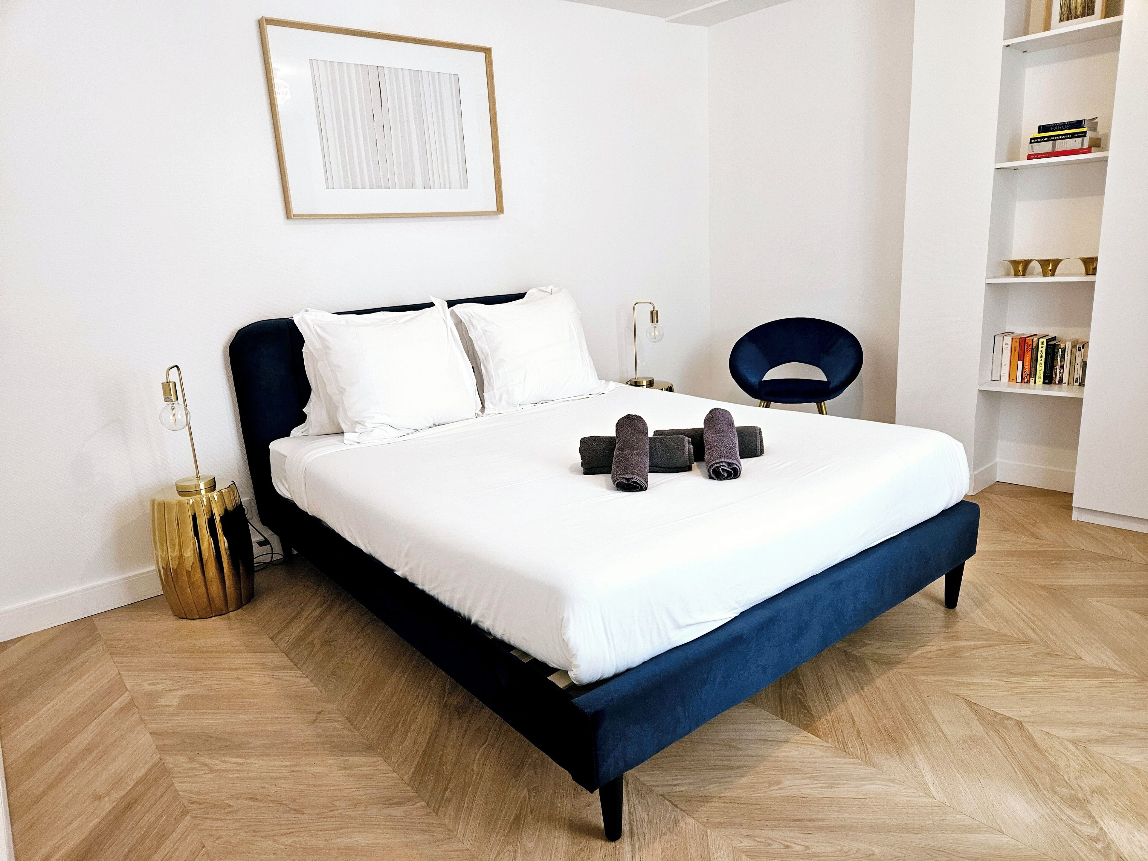 A neatly made bed with white linens, a dark blue headboard, and rolled towels on the bed.