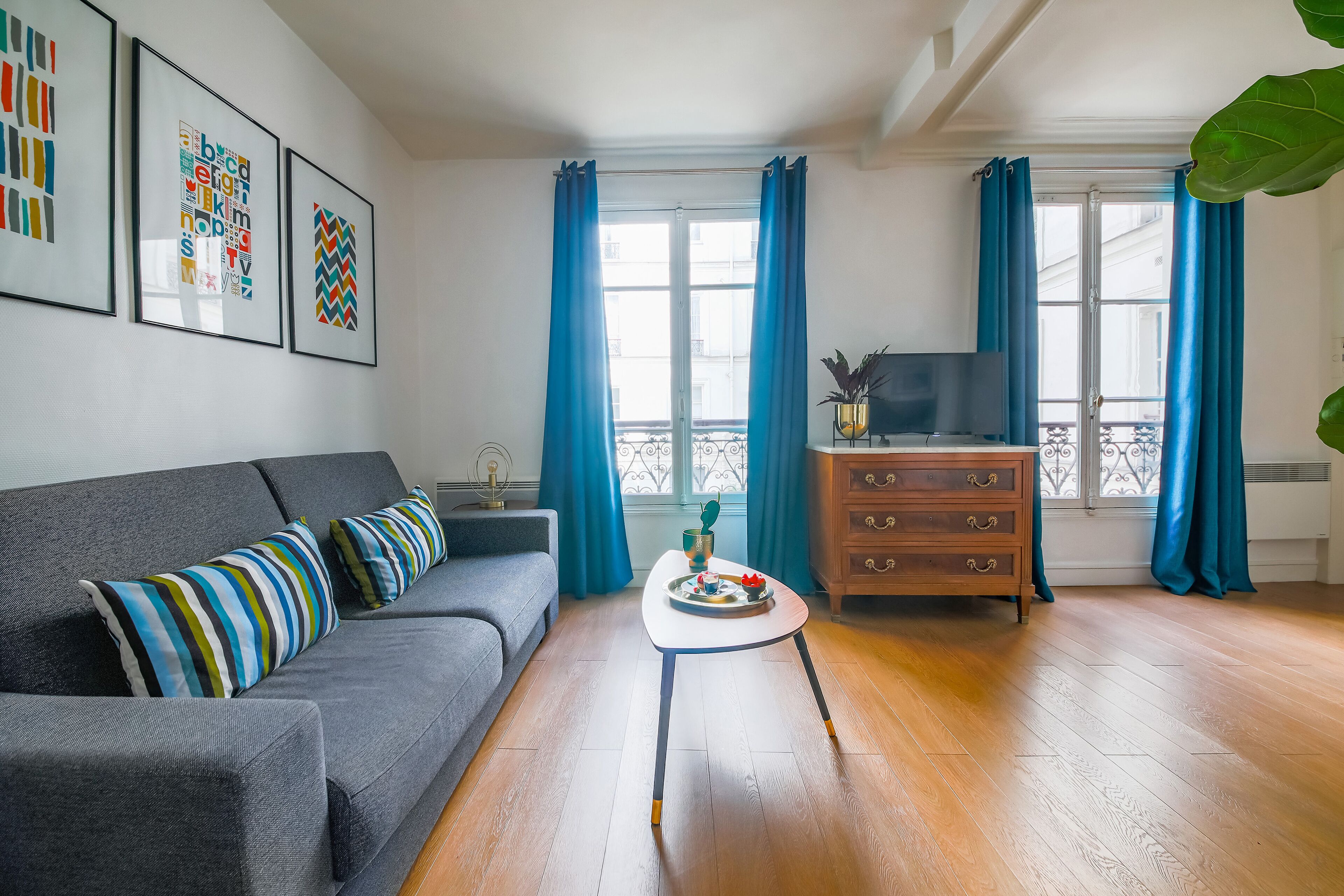 Una sala de estar con un sofá gris, una mesita blanca pequeña, un aparador de madera y un televisor sobre un mueble.