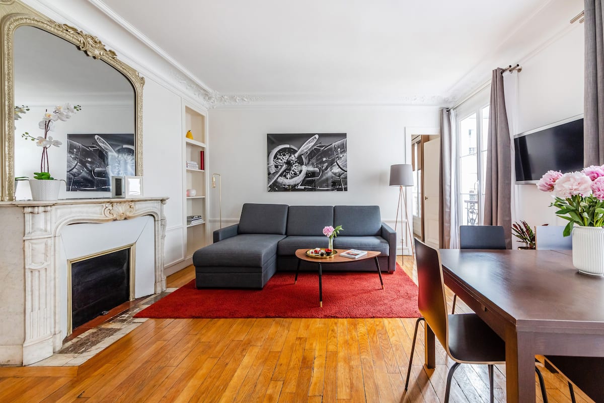 Un salon moderne avec une cheminée, un canapé d’angle gris, un tapis rouge et un coin repas comprenant une table et des chaises en bois.