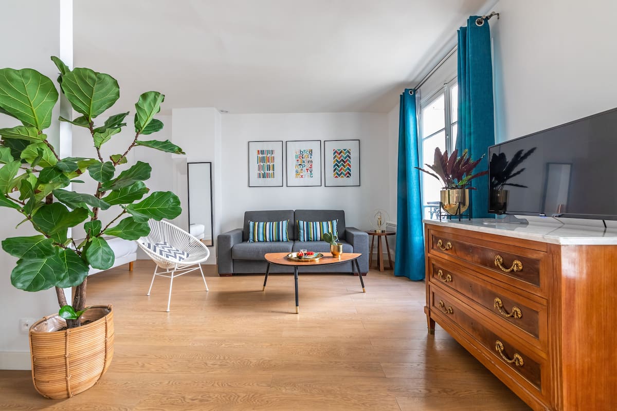 Un salon moderne avec un canapé gris, un fauteuil blanc, une table basse en bois, une grande plante en pot, un meuble en bois et un téléviseur à écran plat.