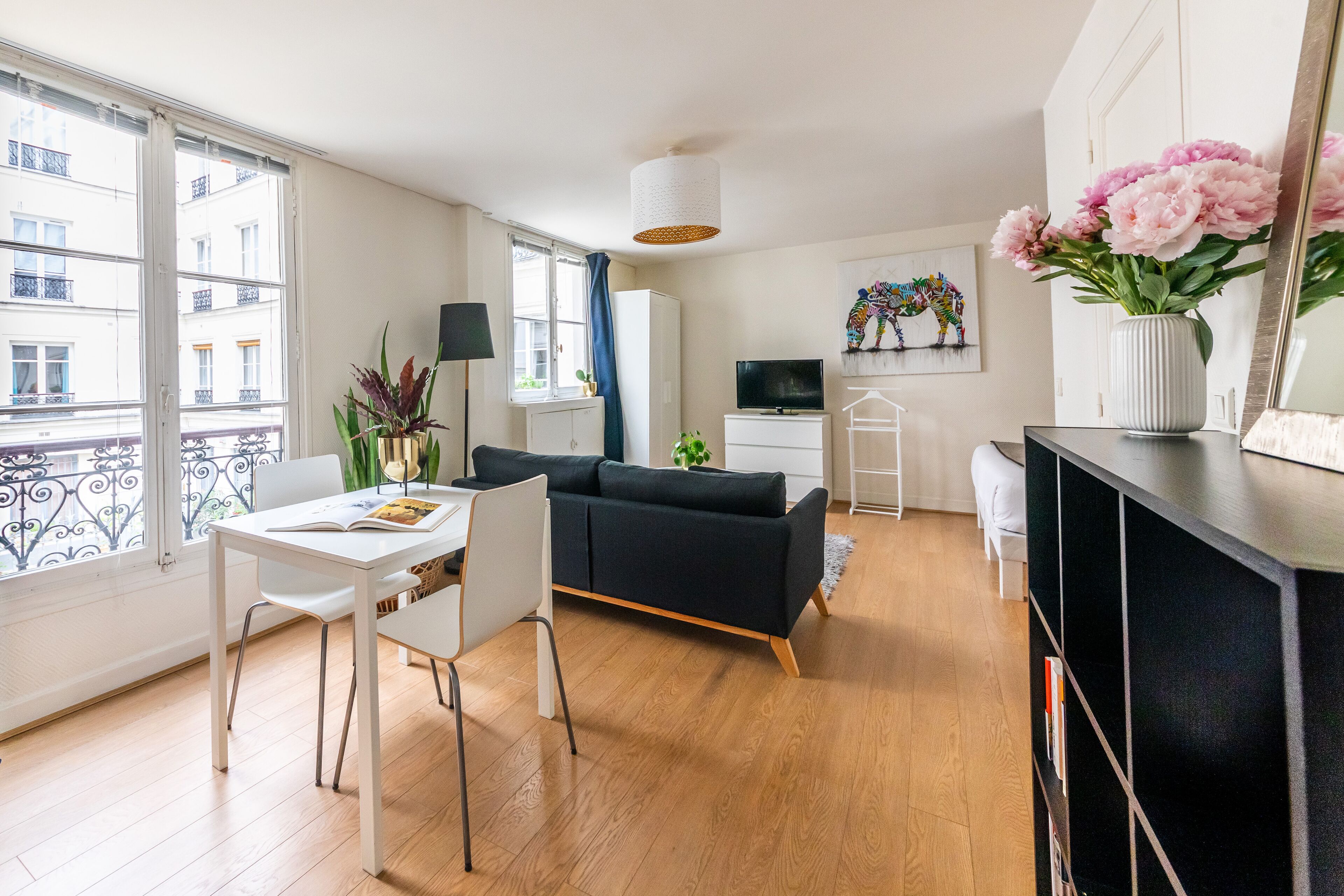 A modern living room with a sofa, dining table, chairs, a TV, and a large window with a view of buildings.