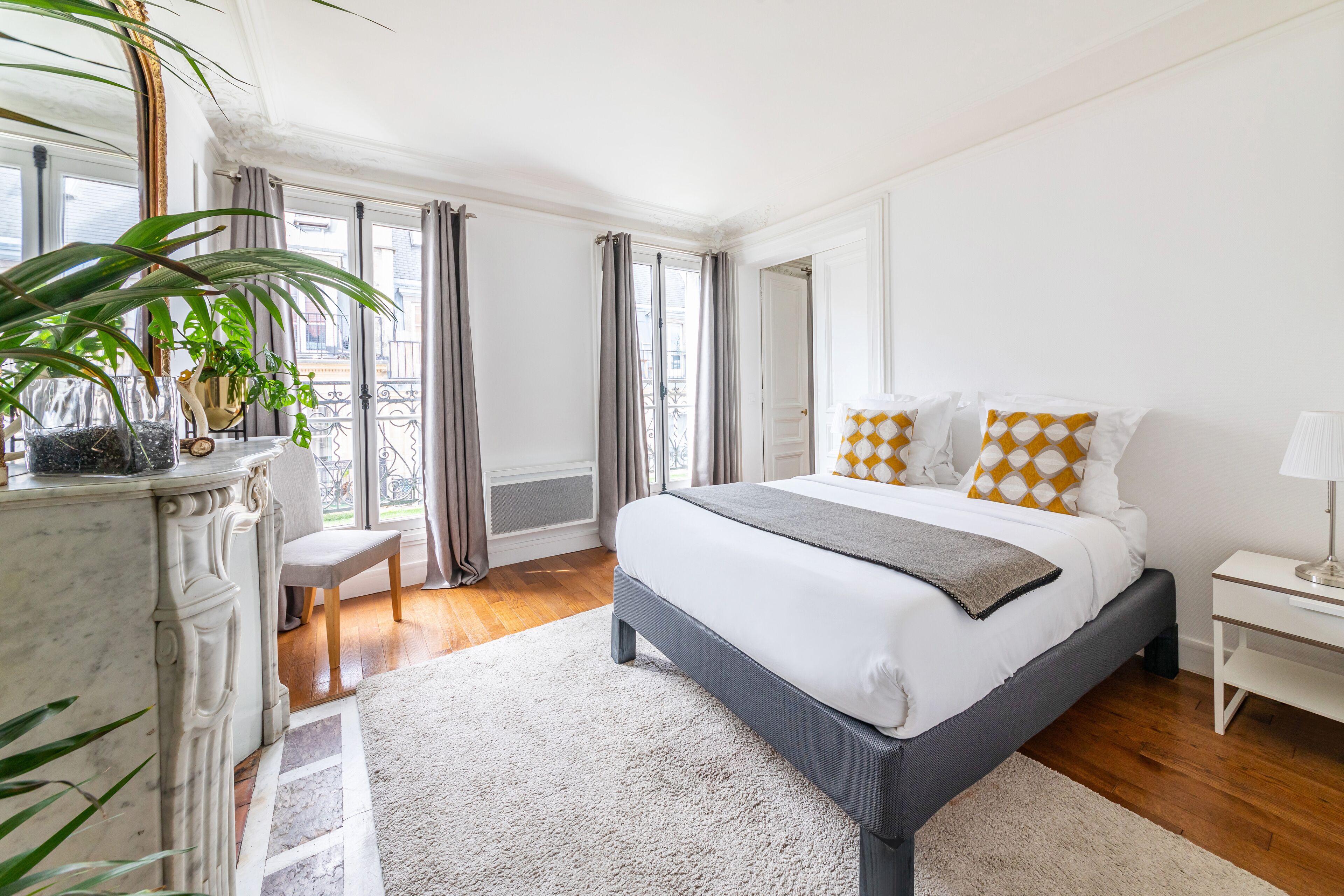 A bedroom with a bed, a nightstand, a lamp, a chair, a potted plant, and a view of the outside.