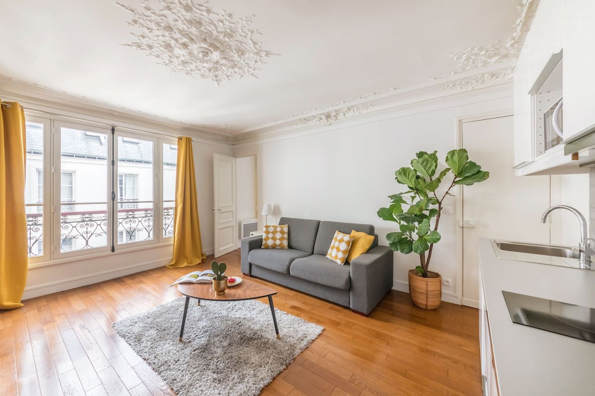 Un salon moderne avec un canapé gris, un tapis blanc, une grande plante en pot et une vue sur la ville grâce à de grandes fenêtres.