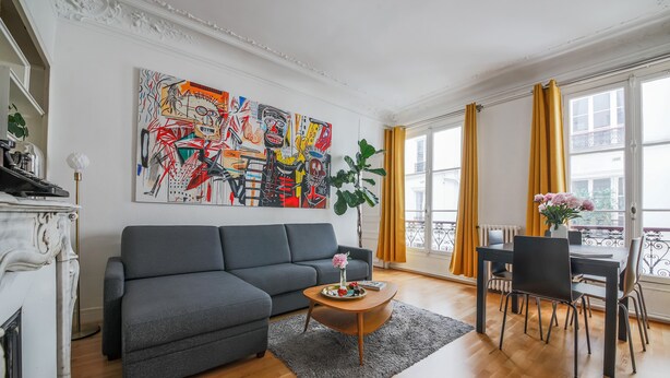 A modern living room with a grey sofa, a wooden coffee table with flowers, a large abstract painting, and a dining area with yellow curtains.