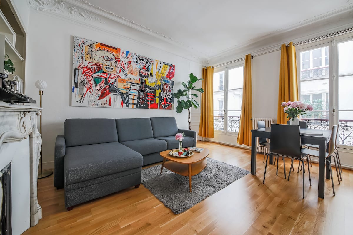 Un salon moderne avec un canapé gris, une table basse en bois agrémentée de fleurs, un grand tableau abstrait et un coin repas avec des rideaux jaunes.