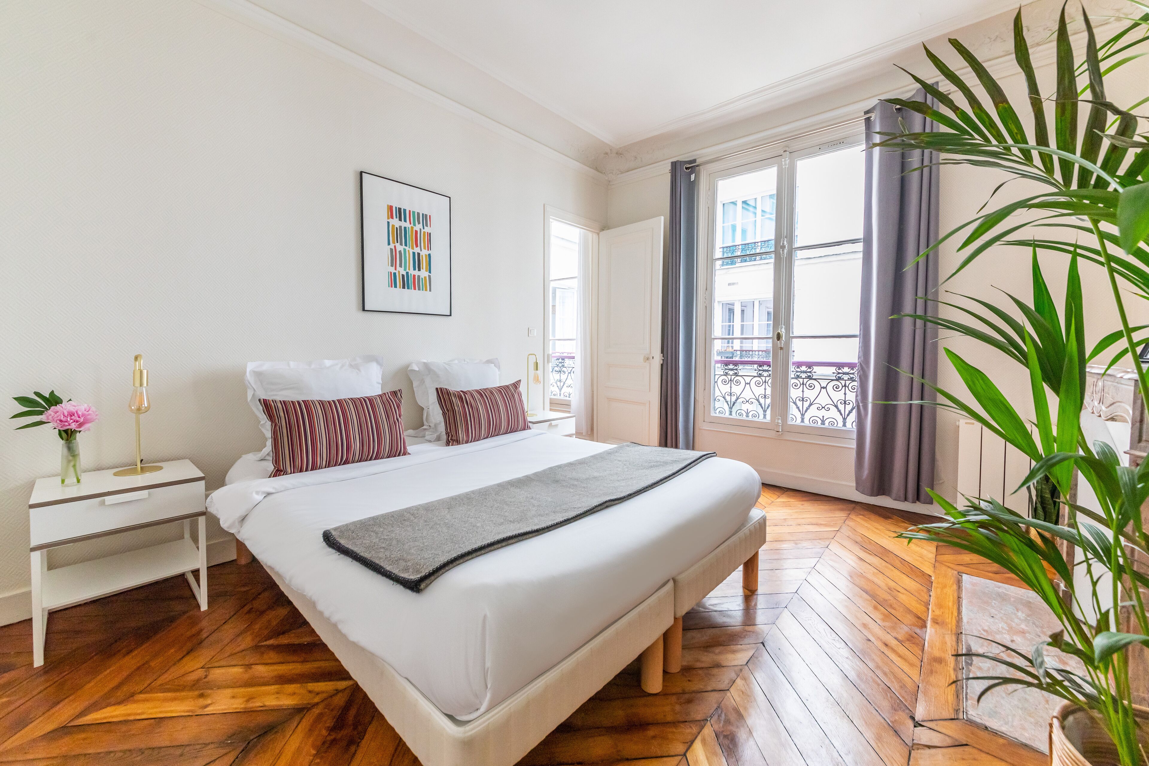 A hotel room with a large bed, a bedside table with a lamp, a vase with flowers, and a window with curtains.