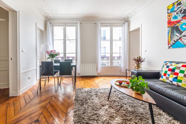 A modern living room with a herringbone wooden floor, a gray sofa, a coffee table with a plant, and a colorful abstract painting on the wall. - Résidence Bergère - Appartements (Paris)