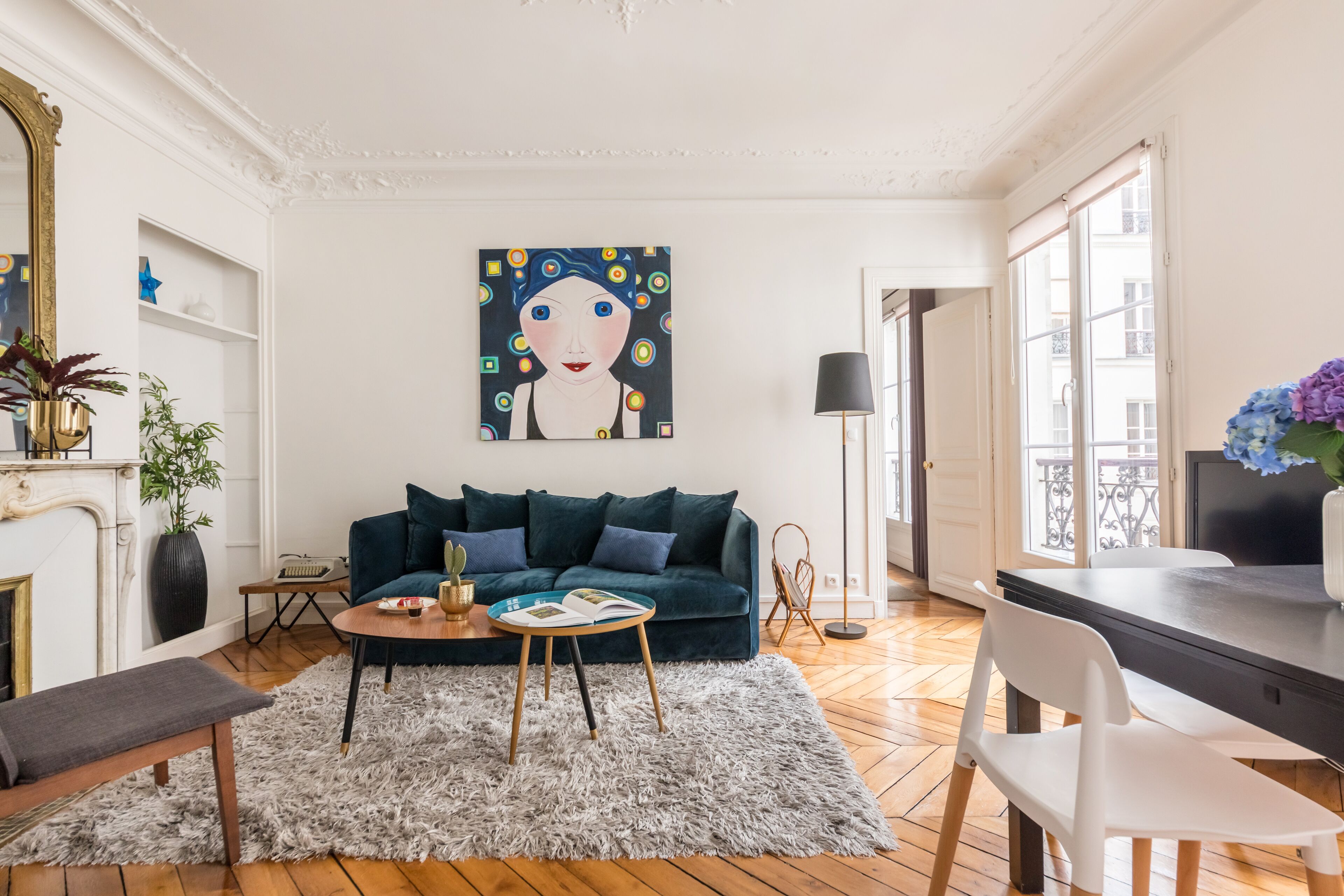 A living room with a dark green sofa, a round coffee table, and a fireplace. There is a painting on the wall and a large window with a view of buildings.