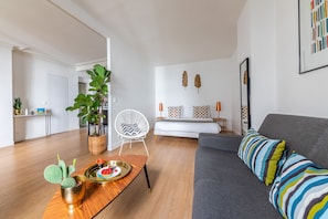 A modern living room with a grey sofa, a wooden coffee table with a cactus, and a white chair. A bed with patterned pillows is visible in the background. - Résidence Bergère - Appartements (Paris)