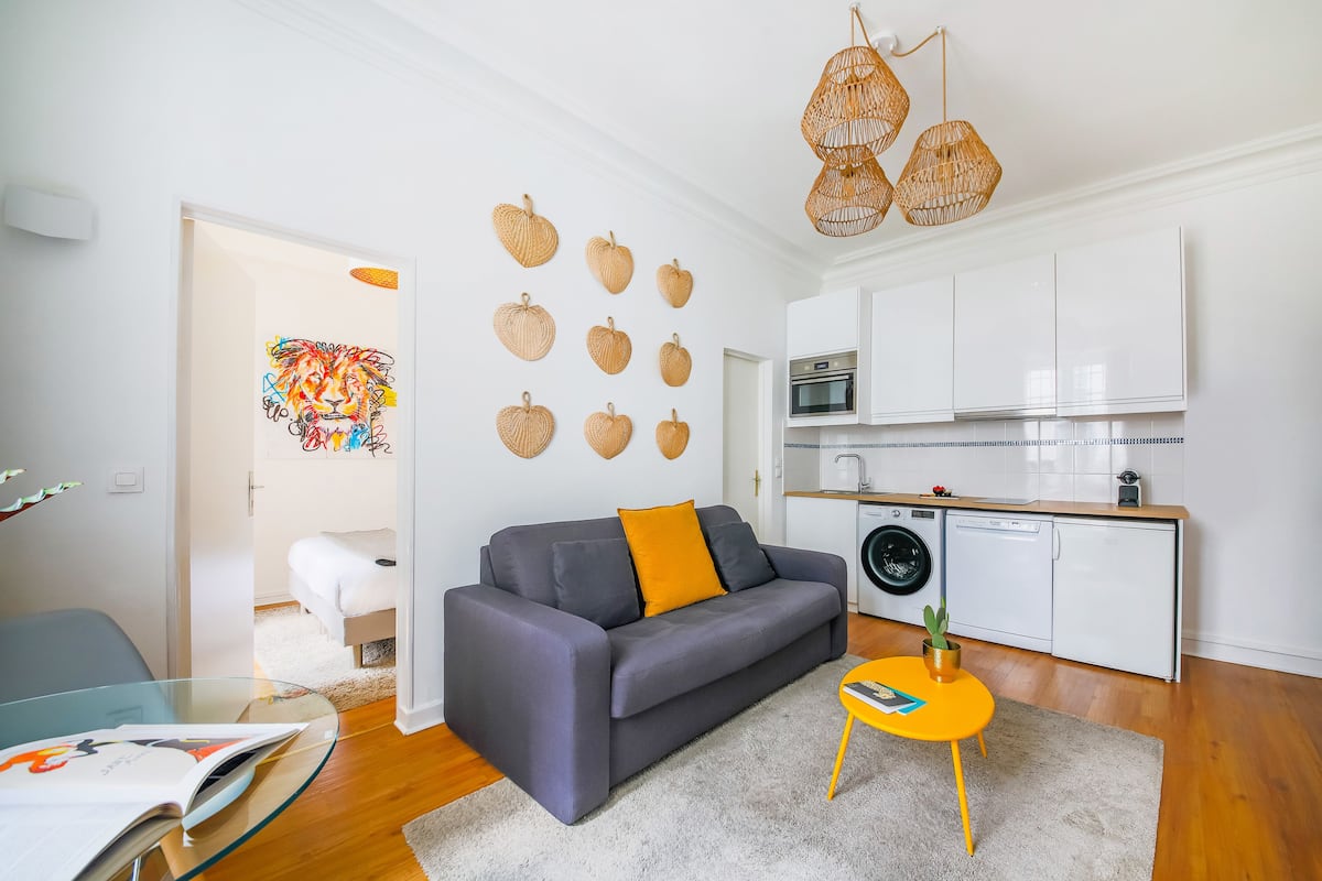 Un salon moderne avec un canapé gris, une petite table jaune et un coin cuisine doté de meubles blancs et d’un lave-linge.