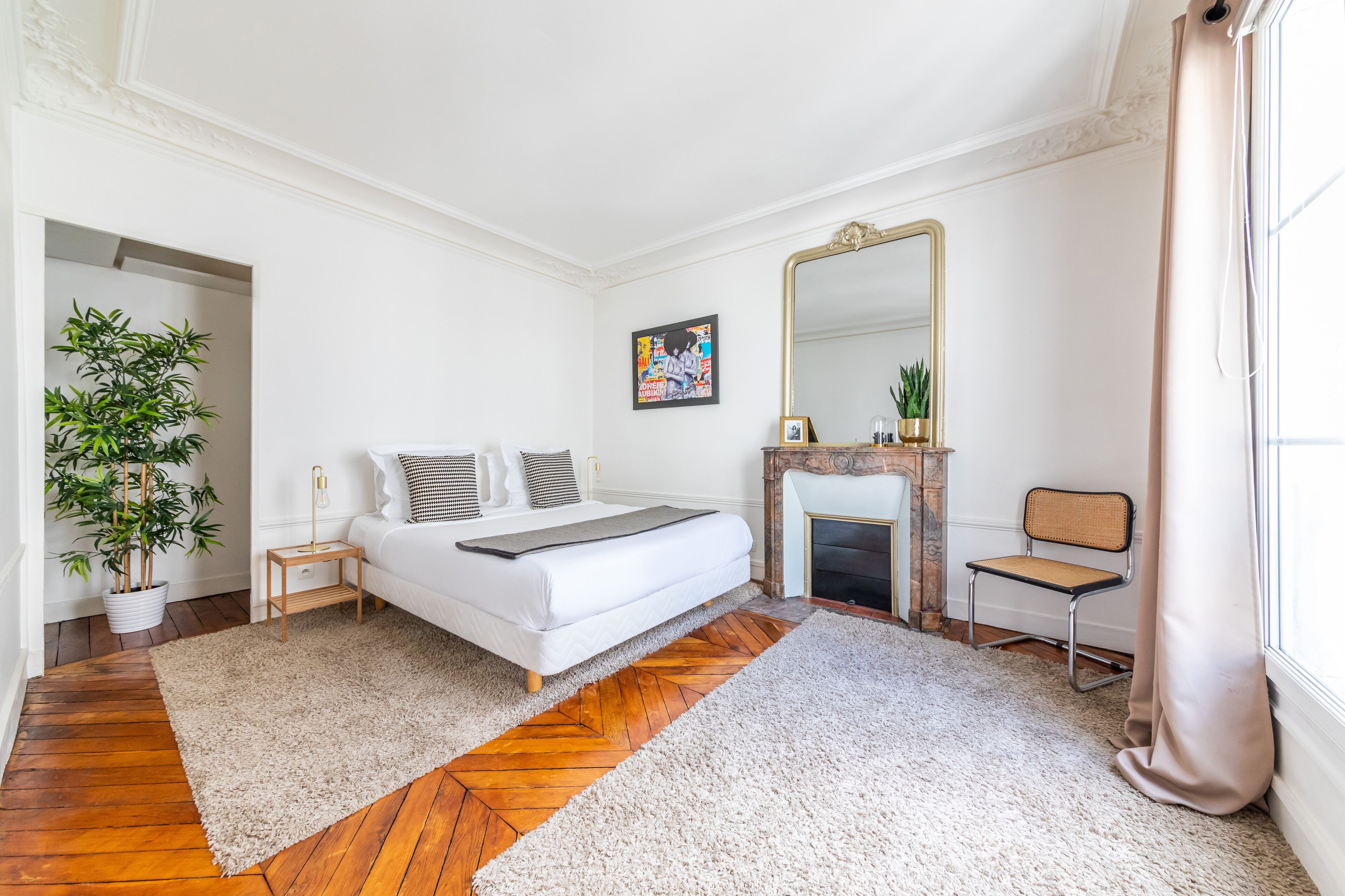 A bedroom with a bed, a mirror, a fireplace, a chair, and a potted plant.