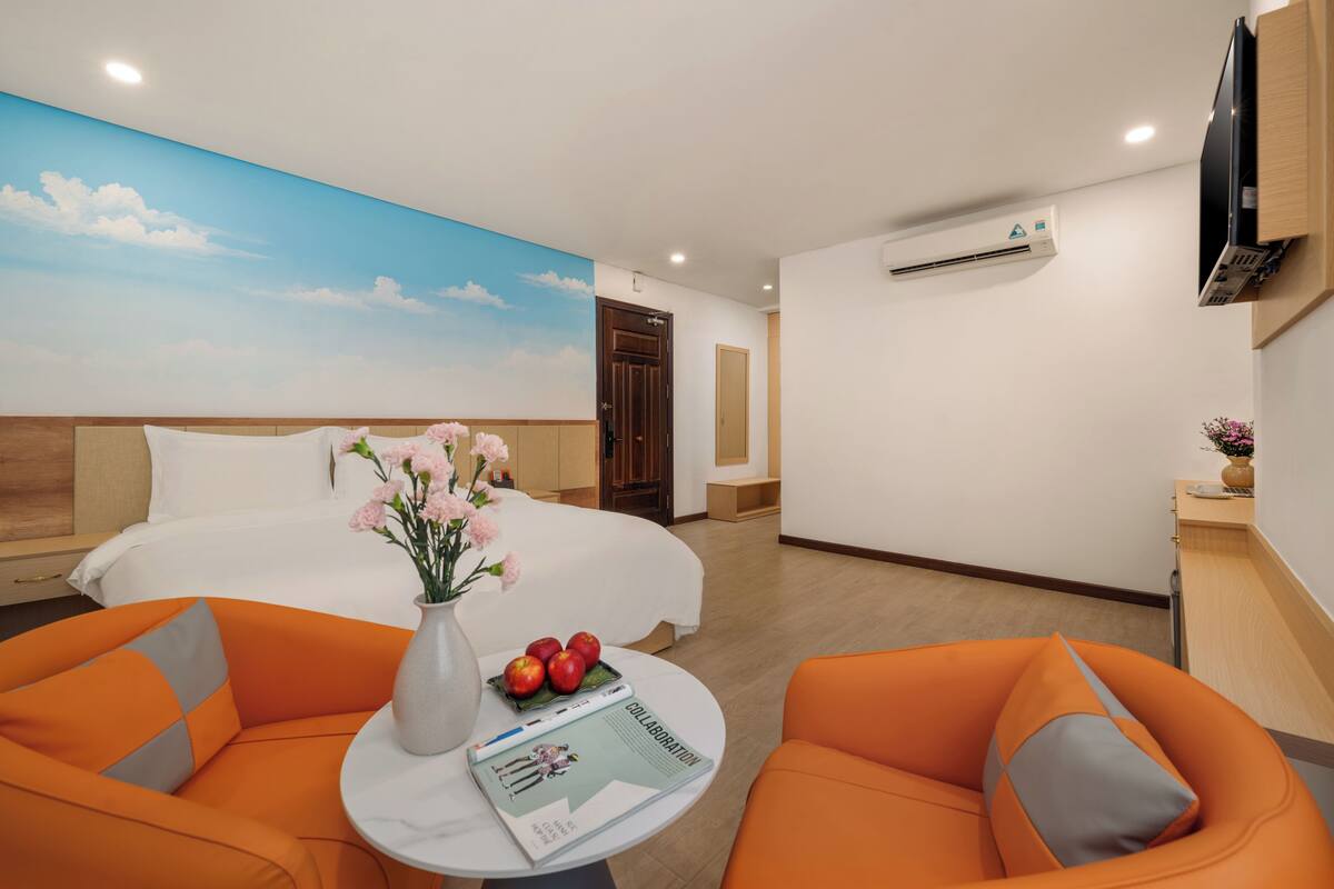 A hotel room with a bed, two orange chairs, a small table with a vase of flowers and apples, and a wall-mounted TV.