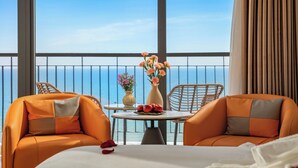 A hotel room with a balcony, two orange armchairs, a small round table with a vase of flowers, and a bed with white linens. - Lunar Hotel Danang (Da Nang)