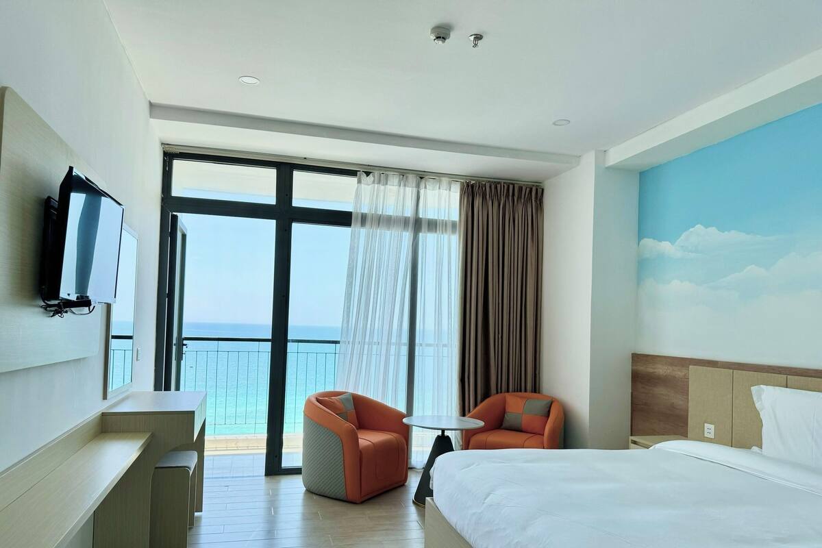A hotel room with a bed, two armchairs, a desk, a TV, and a large window with a view of the sea.