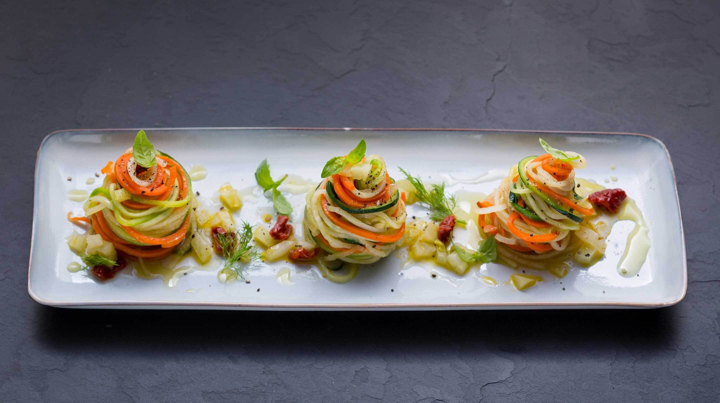 A rectangular plate with three spiral vegetable dishes, garnished with herbs, on a dark surface.
