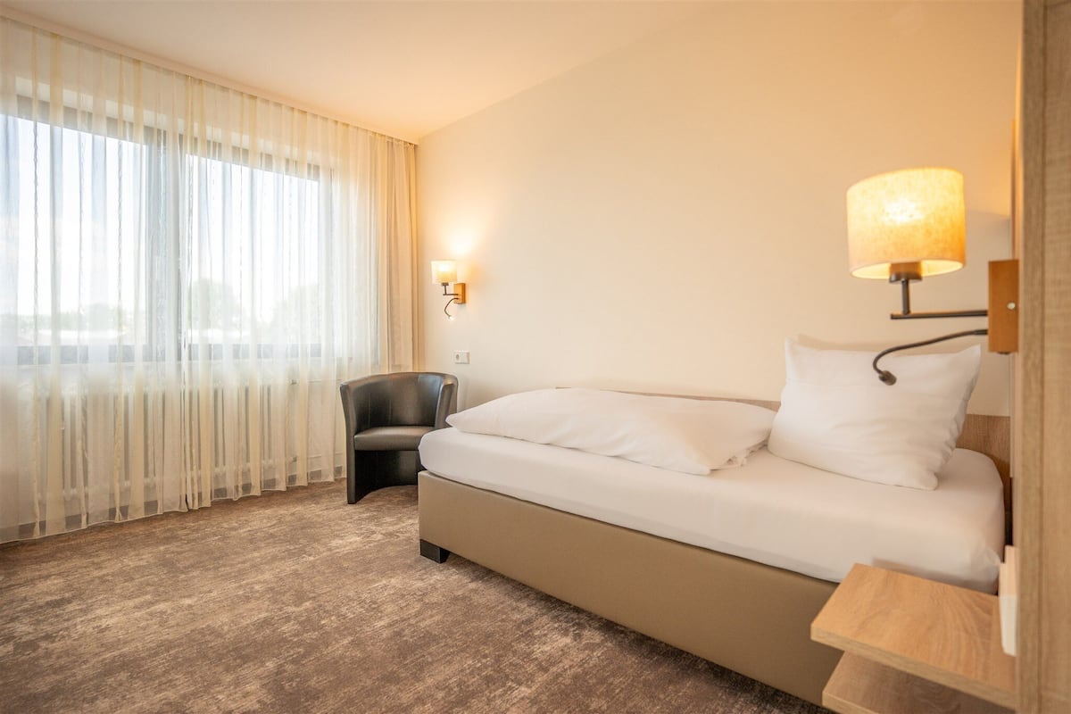 Une chambre d’hôtel avec un lit, une chaise, une lampe et une fenêtre avec des rideaux légers.