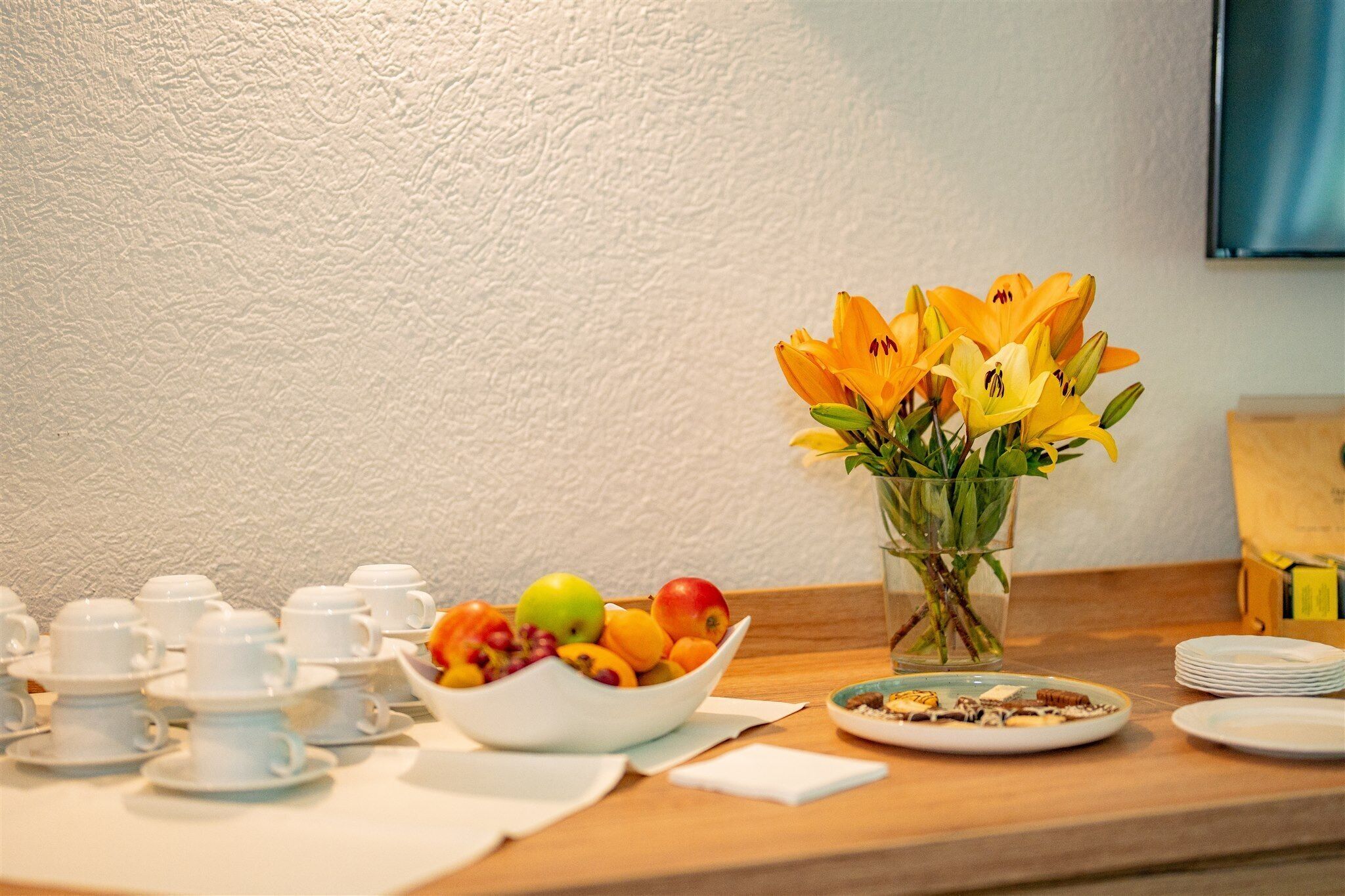 A breakfast setup with a fruit bowl, a vase of flowers, and tea cups on a wooden table.