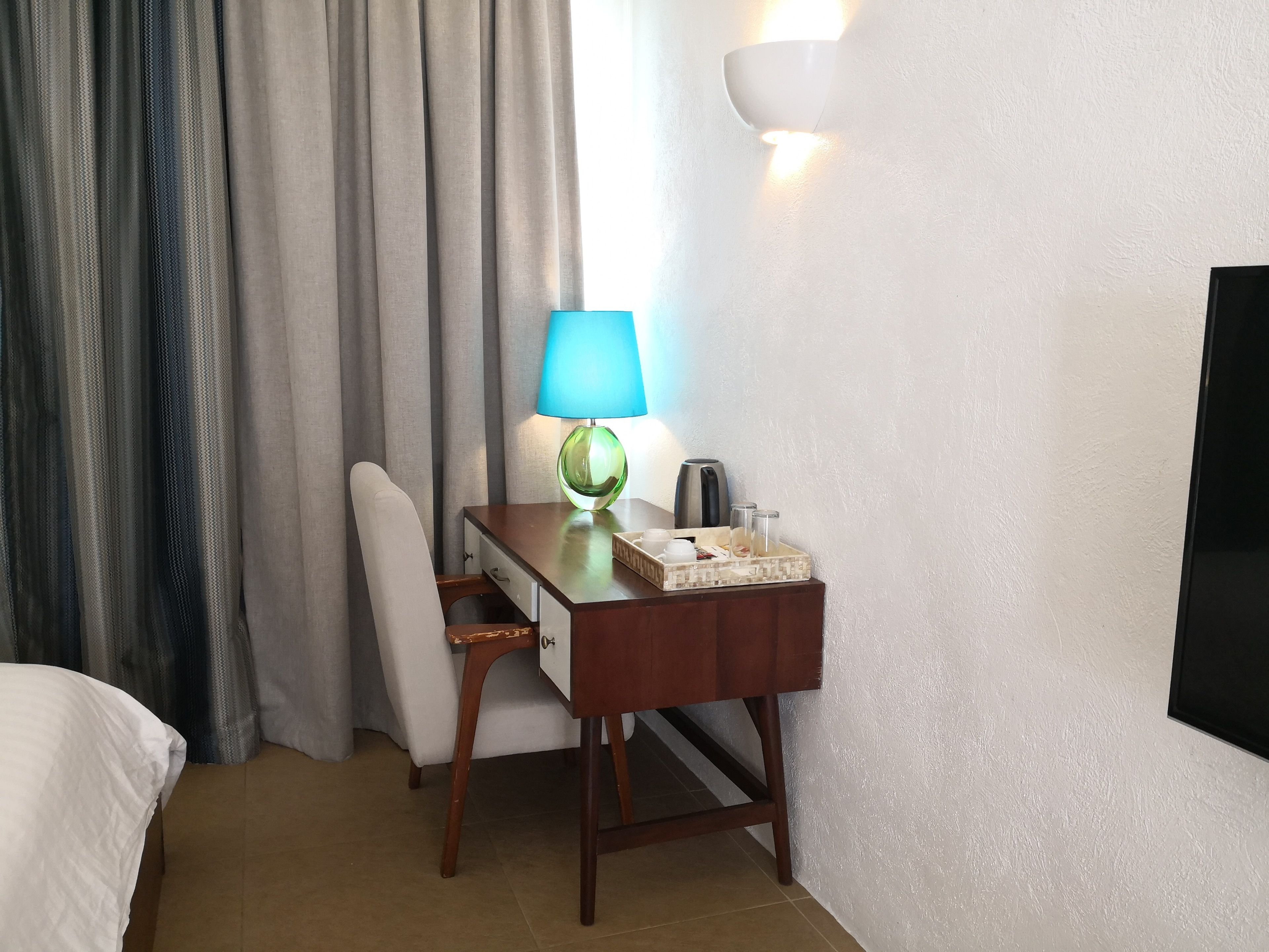 A hotel room with a wooden desk, a chair, a lamp, a tray with cups and a box, and a TV mounted on the wall.