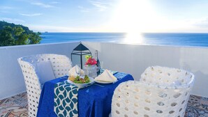 Una mesa con mantel azul, sillas blancas, una botella de agua, un vaso y una vela en un balcón con vista al océano.