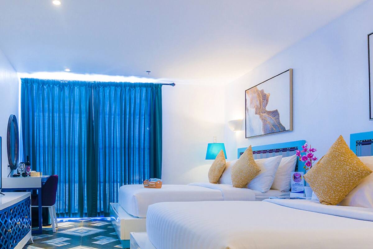 Habitación de hotel con dos camas, un escritorio con una computadora, un televisor y vistas al mar a través de cortinas azules.