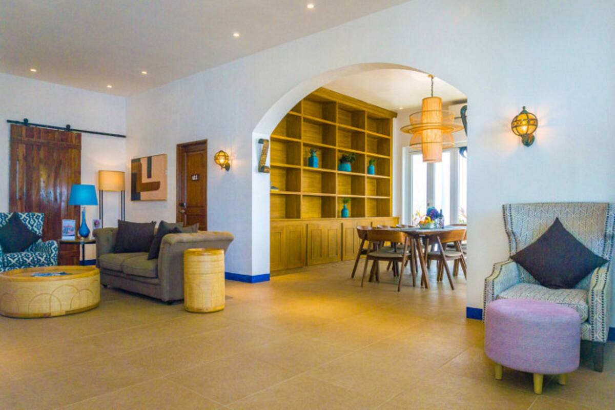 Amplia sala de estar con sofá, reposapiés y sillón, una mesa de comedor con sillas, zona de cocina con armarios de madera y un amplio arco de entrada.