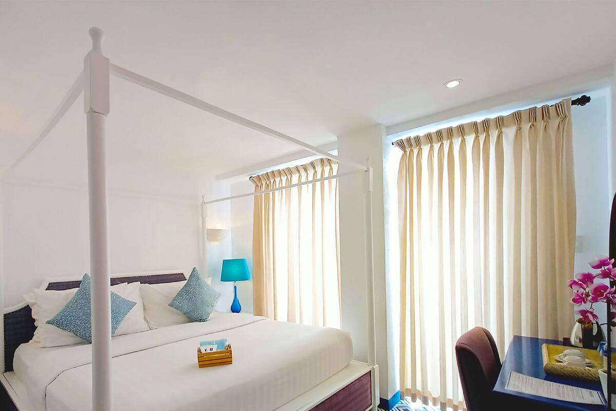 Una habitación de hotel con cama con dosel, dos lámparas de noche, una mesita de noche, un escritorio con silla y una ventana con cortinas.