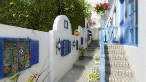 Un estrecho pasillo de paredes blancas con contraventanas y puertas azules, que conduce a un edificio de fachada de piedra.