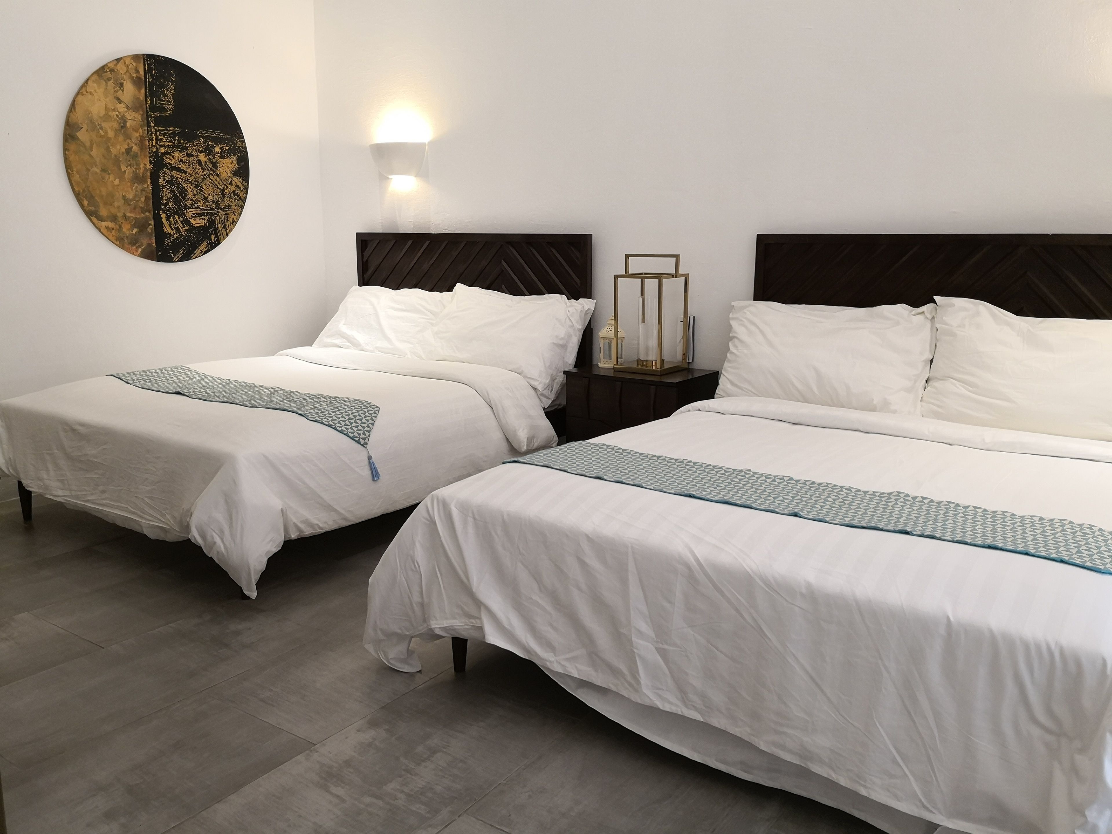 A hotel room with two beds, a nightstand with a lamp, and a wall-mounted light fixture.
