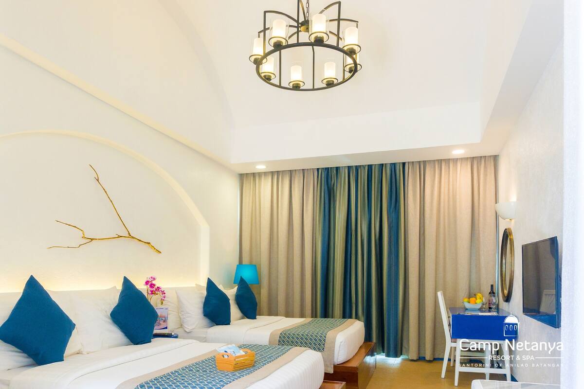 Habitación de hotel con dos camas, un candelabro, un escritorio con silla, un televisor y un ventanal con cortinas.