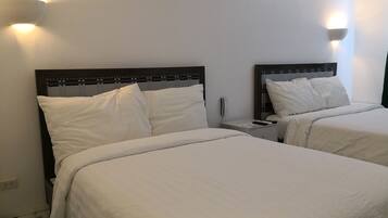 Dos camas con ropa blanca, una mesita de noche con teléfono y control remoto, y lámparas fijadas en la pared.