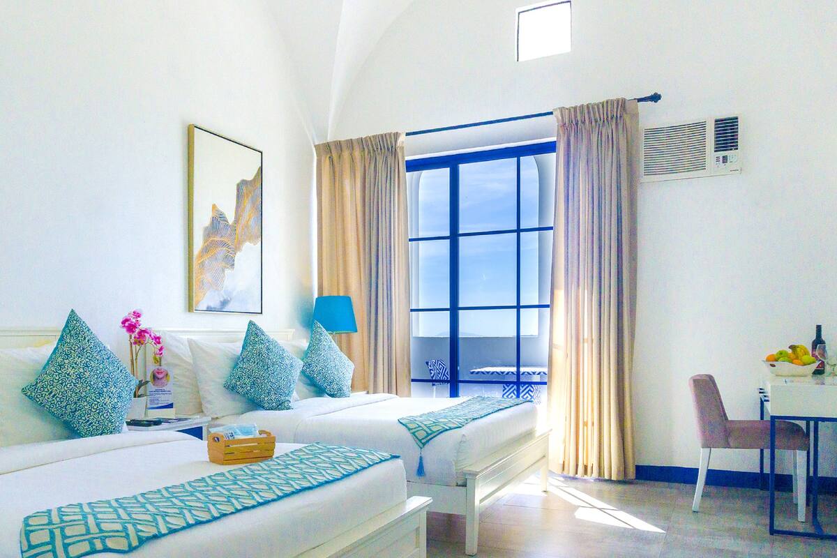 Una habitación de hotel con cama, escritorio con silla, ventana con cortinas y obras de arte en las paredes.